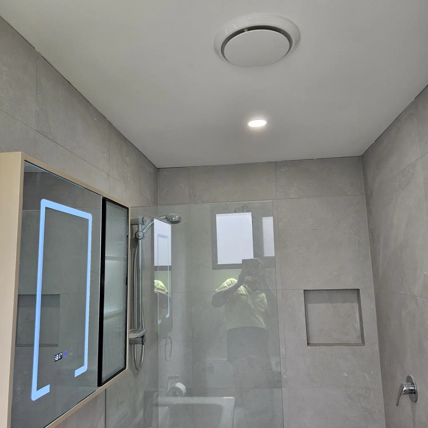 Grey-tiled bathroom with backlit LED mirror cabinet, ceiling exhaust fan and downlight over the shower