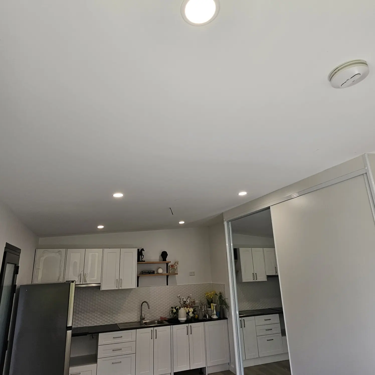 Galley kitchen with white cabinetry lit by recessed LED downlights and an oyster light in the hallway