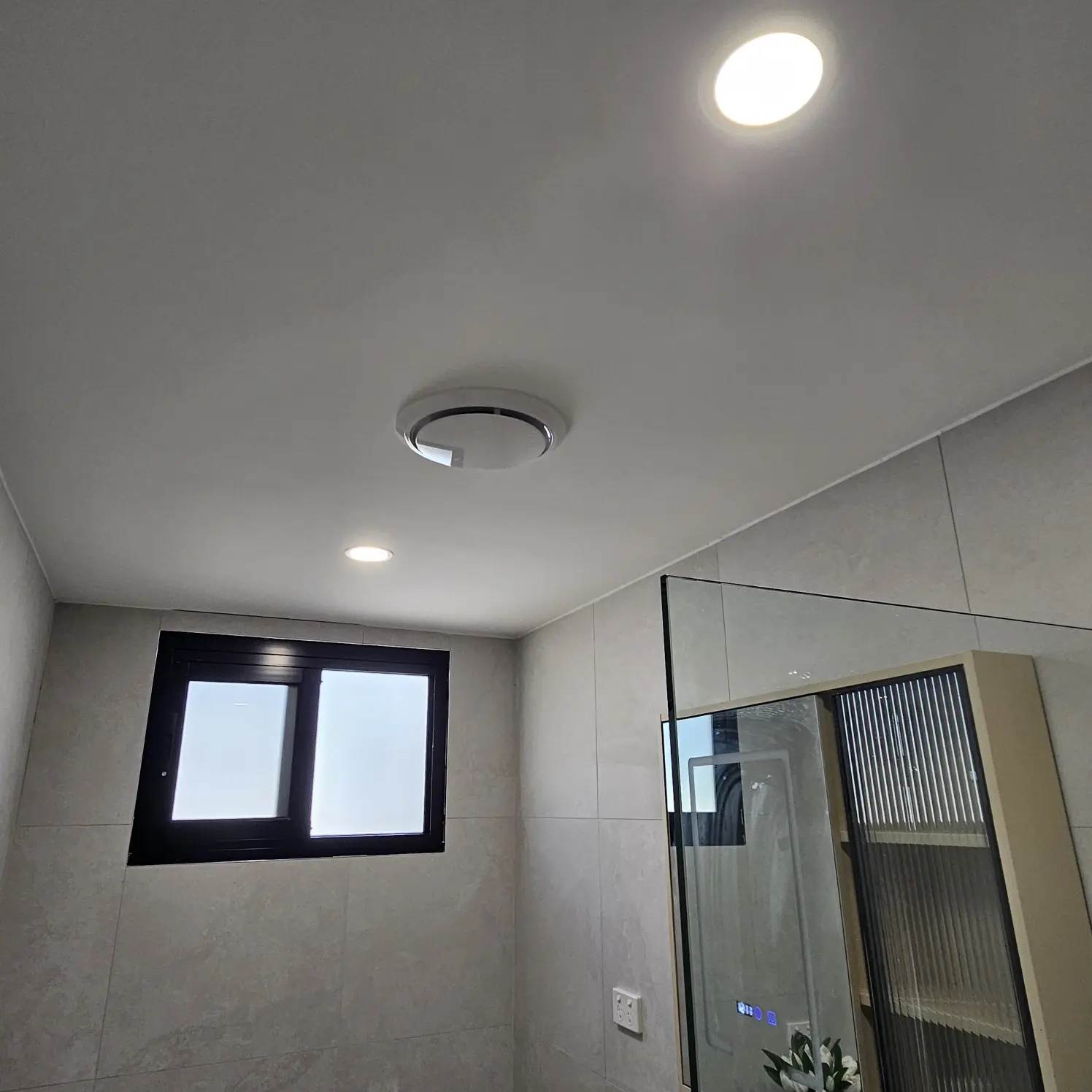 Tiled bathroom with a round LED oyster ceiling light, downlight and exhaust fan above the shower screen